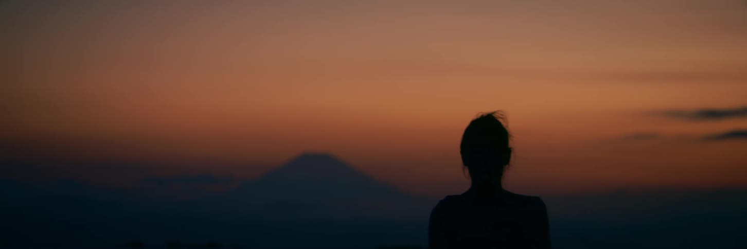 A silhouette of a contemplative woman with her hair blowing in the breeze stands against a beautiful sunset with a mountain in the distance
