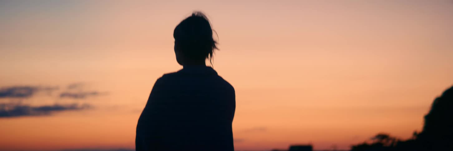 A soft-focus silhouette of a woman with her hair in a bun peacefully watching the warm sunset sky