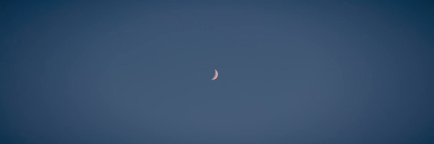 A lone crescent moon sits peacefully in the center of a clear, dark blue sky at twilight