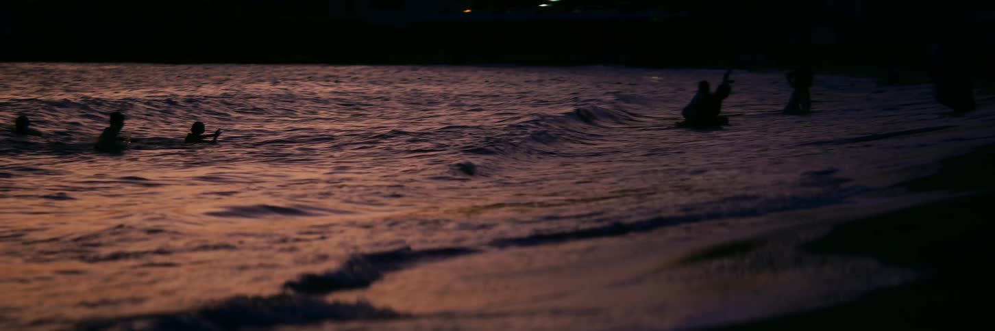 Silhouetted figures enjoy a late evening swim in the ocean as gentle waves roll ashore in slow motion