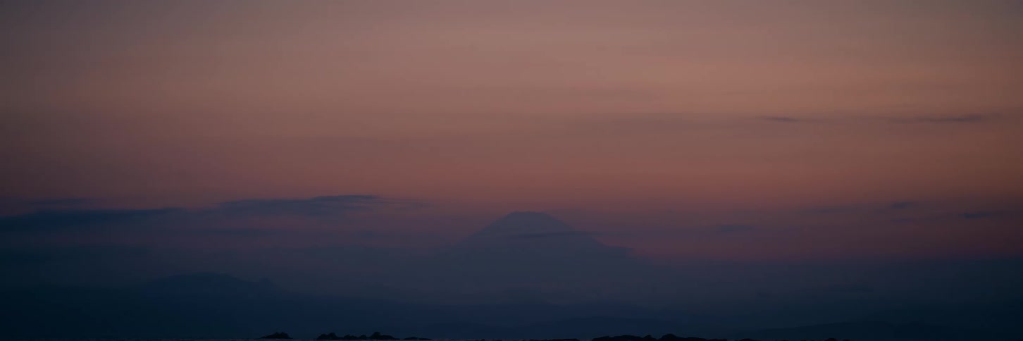 The silhouette of Mount Fuji is seen in the distance against a hazy purple and pink sky at dusk