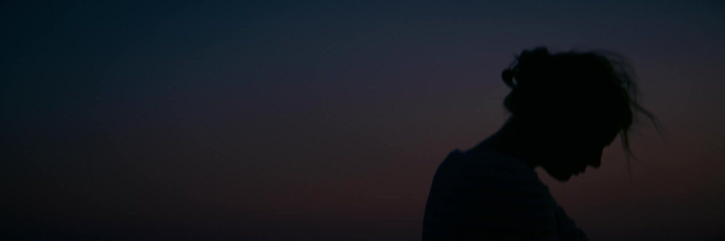 A lonely silhouette of a woman from behind contemplates her sadness against a darkening twilight sky