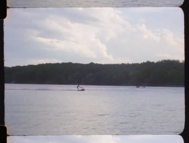 Vintage Super 8 footage captures a person riding a personal watercraft across a calm lake on an overcast day