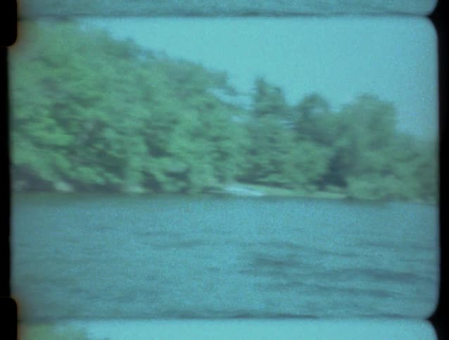 Vintage 8mm footage of a boat ride on a lake, passing a forested shoreline and a pontoon boat