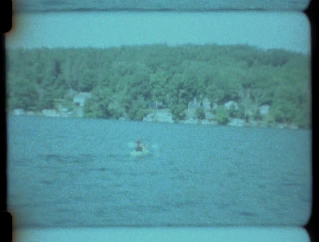 A person paddles a kayak across a vast lake with a tree-lined shore in this grainy, vintage 8mm film footage