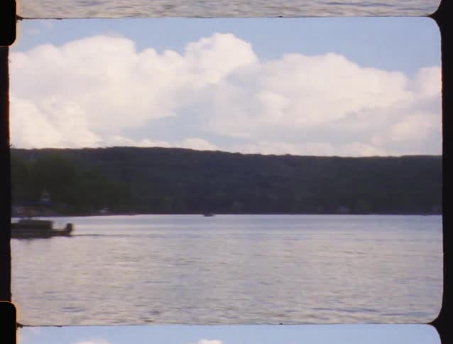 Vintage 8mm film pans across a scenic lake, showing boats cruising on the water under a sky that slowly fills with storm clouds