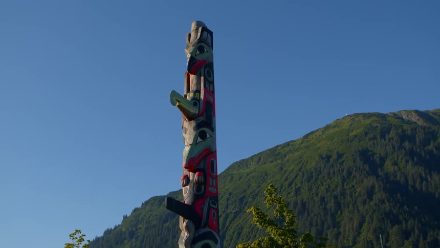 A colorful Indigenous totem pole stands tall with a lush green mountain and clear blue sky in the background