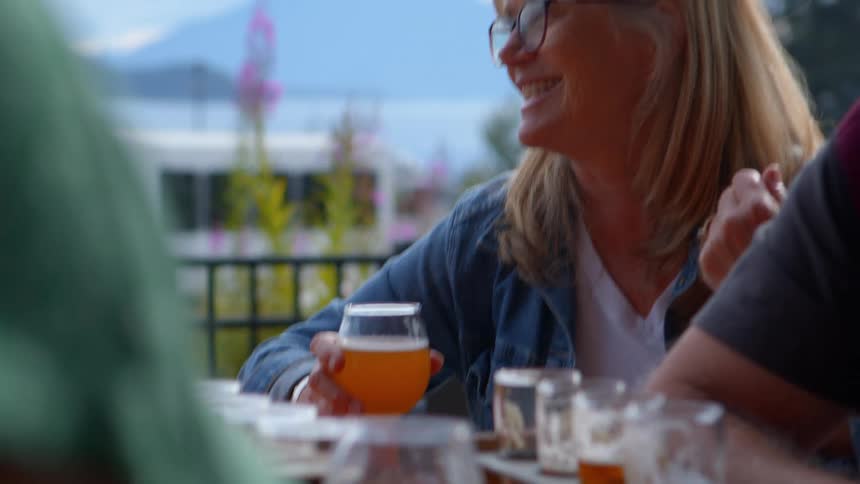 A happy blonde woman with glasses laughs and socializes with friends while enjoying a beer flight on an outdoor patio