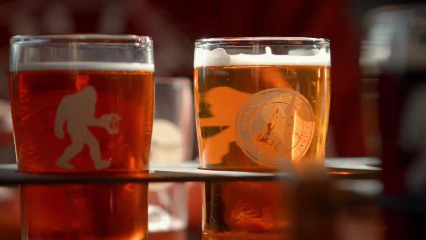 Golden craft beer tasters with Sasquatch logos sit on a flight paddle in a sunlit brewery in slow motion
