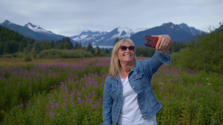 A happy mature woman in sunglasses and a denim jacket takes a selfie in a field of purple wildflowers with a scenic mountain range in the background
