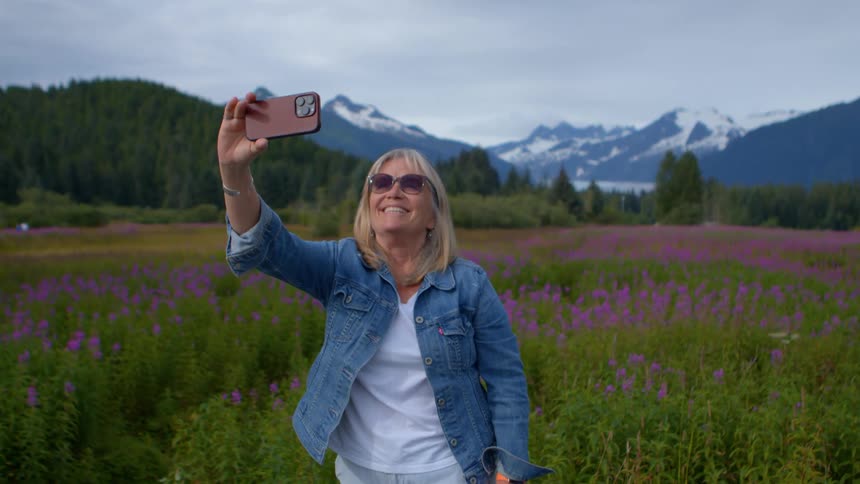 A happy woman takes a selfie video while standing in a scenic field of purple wildflowers with snow-capped mountains in the background