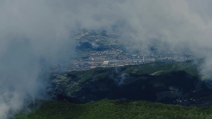 Wispy clouds slowly part to reveal an industrial town and factory nestled in a green mountain valley
