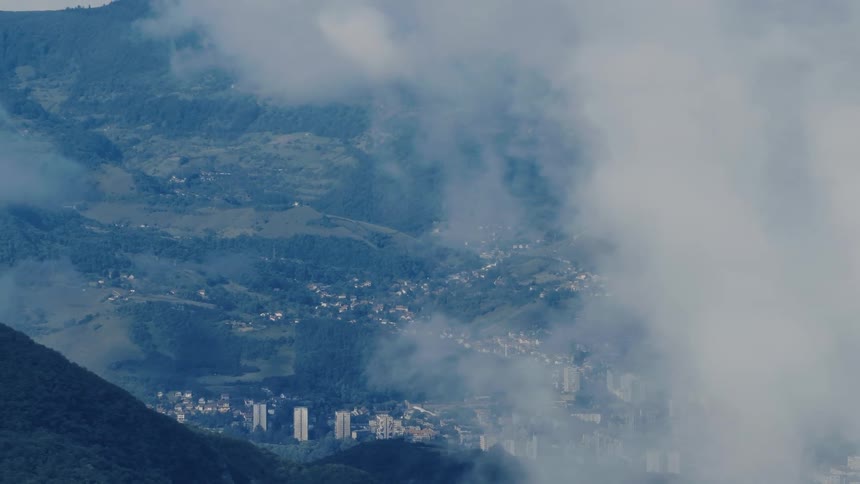 An aerial view reveals a city nestled in a lush green valley through a veil of slow-moving clouds