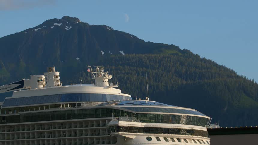 The top of a large cruise ship sits peacefully in port with a stunning view of a forested mountain and the daytime moon