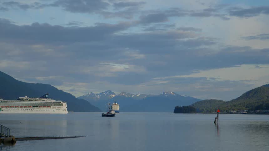A serene view of a bay with a fishing boat sailing past cruise ships, set against a backdrop of stunning mountains and a cloudy sky