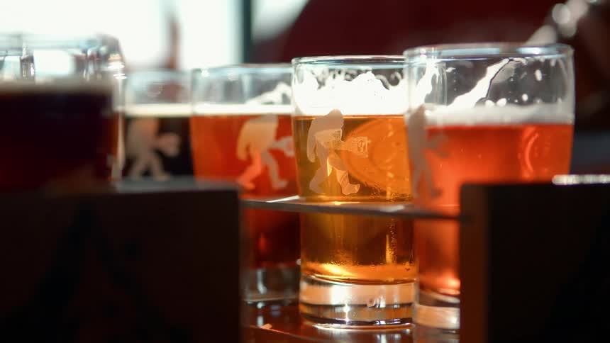 A flight of craft beers with a Bigfoot logo on the glasses sits on a wooden holder in the sunlight