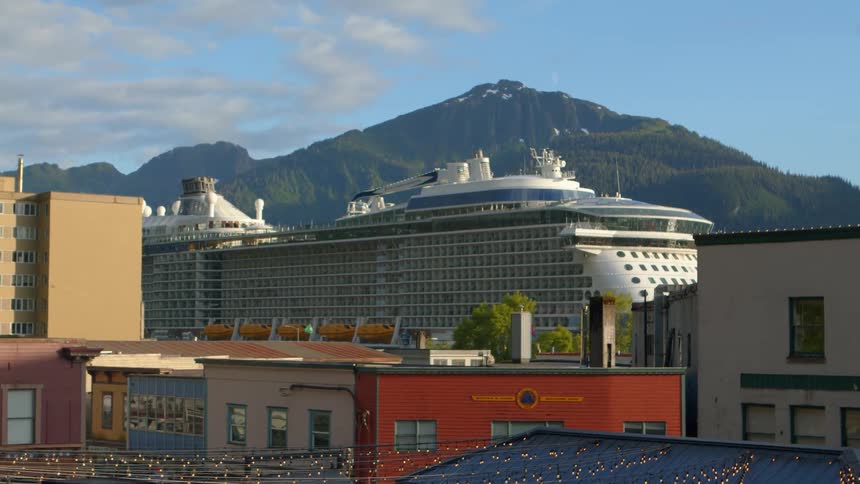 Time-lapse of a massive cruise ship departing from a port city at the base of a majestic mountain during golden hour