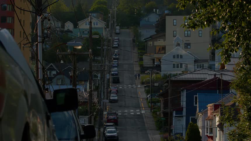 A static long shot captures a steep residential street in a small town, with cars parked on both sides and people walking in the distance