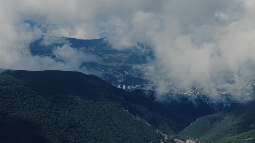 A wide shot of a vast mountain range with low hanging clouds partially obscuring a city in the valley below
