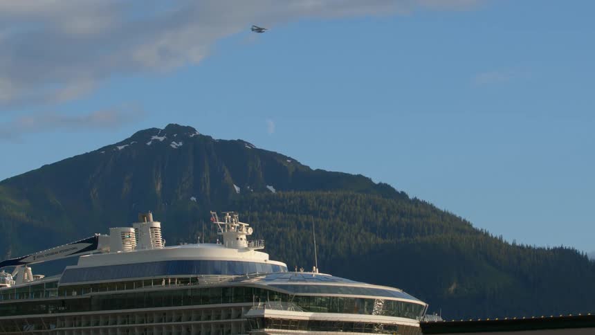 A seaplane flies past a docked cruise ship with a snow-capped mountain and the daytime moon visible in the clear blue sky