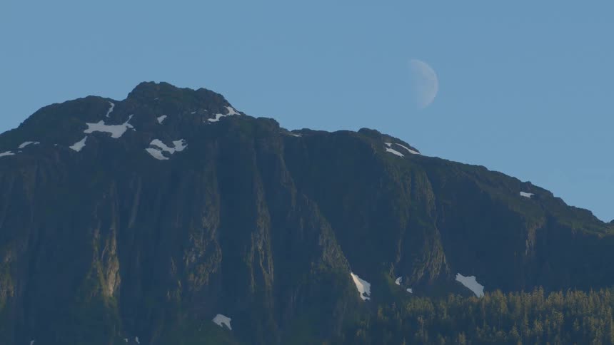 A half-moon is visible in the clear blue sky over a snow-capped mountain peak at dawn