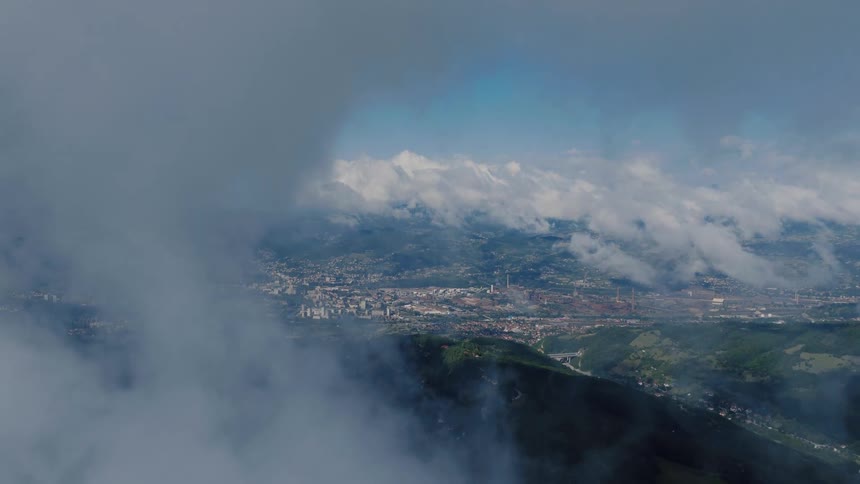 Static aerial shot through wispy clouds showing a distant city and industrial area surrounded by mountains