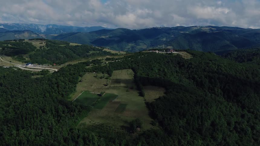 Aerial view of a vast mountain landscape with lush green forests and meadows, as cloud shadows slowly drift across the hills