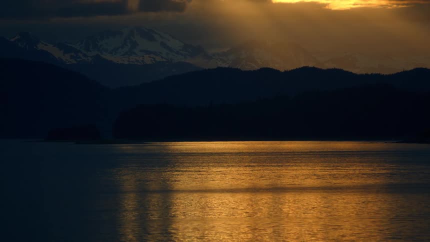 Majestic sunbeams pierce through stormy clouds at sunset, casting a golden reflection on a serene lake surrounded by silhouetted mountains
