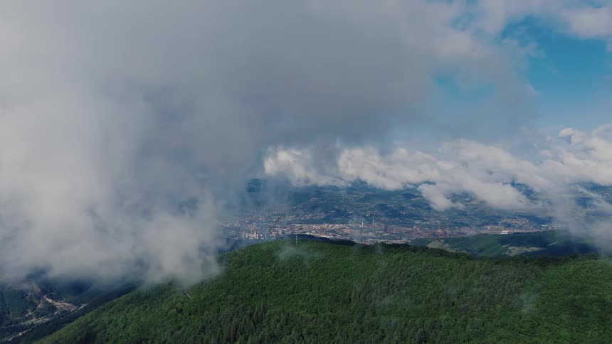 Aerial view from above the clouds reveals a lush green mountain landscape with a distant city nestled in the valley