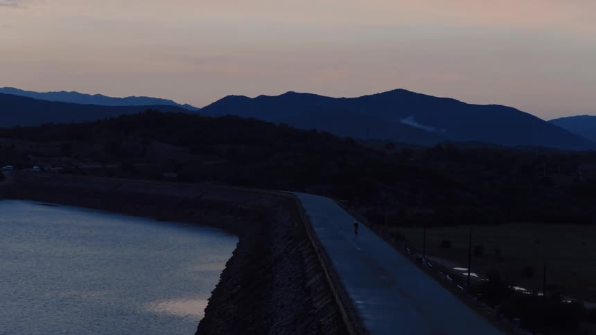 A cyclist rides alone on a dam road into the twilight with a beautiful mountain landscape