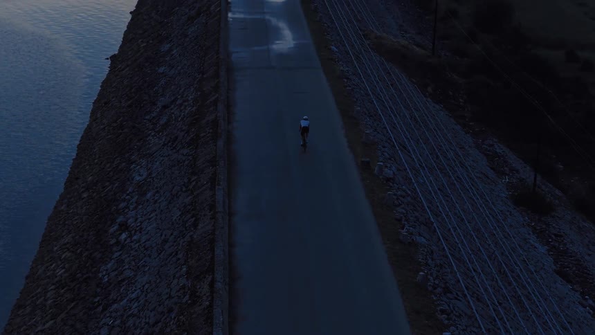 High-angle aerial shot follows a lone cyclist riding along a dam next to a tranquil body of water at twilight