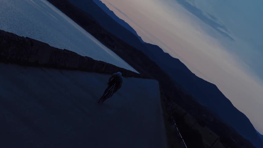 A vertical, moody shot of a cyclist in silhouette riding along a mountain lake path during a beautiful twilight evening