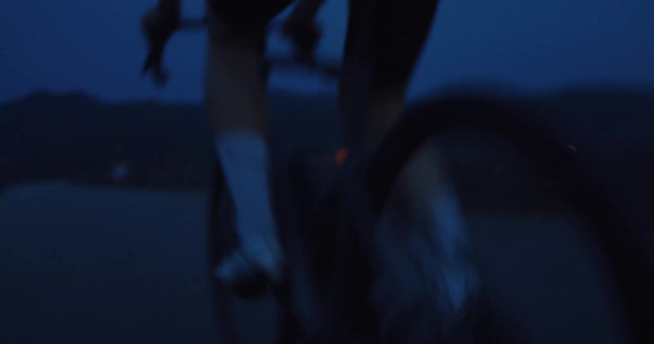 A blurry, fast-paced close-up of a cyclist pedaling on a road bike in the dark blue light of dusk