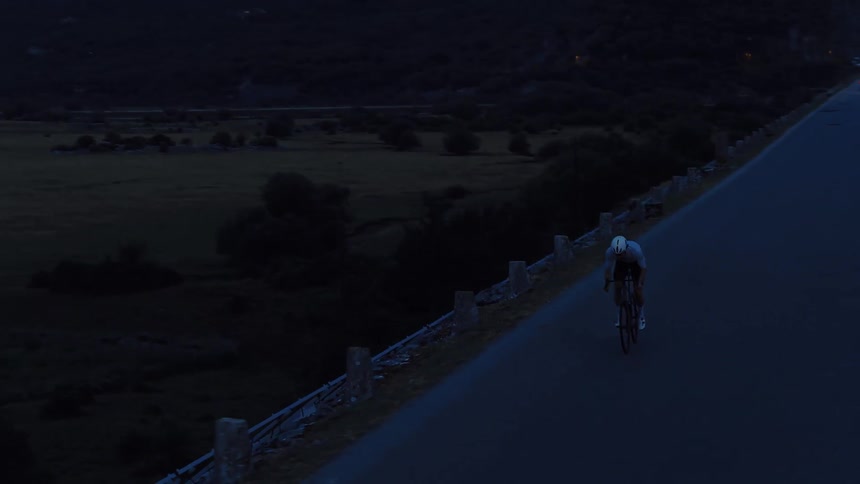 Cinematic aerial shot follows a solitary cyclist training on a dark road over a dam at dusk
