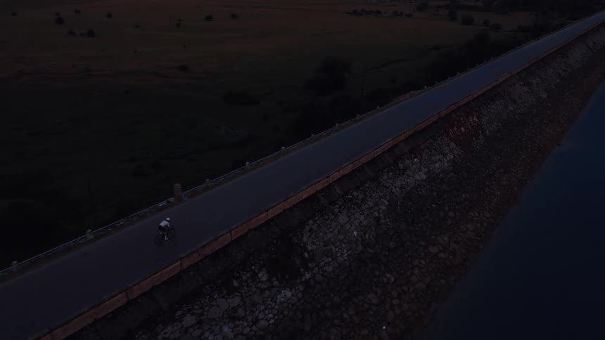 High angle tracking shot of a cyclist riding on an empty road over a dam at dawn