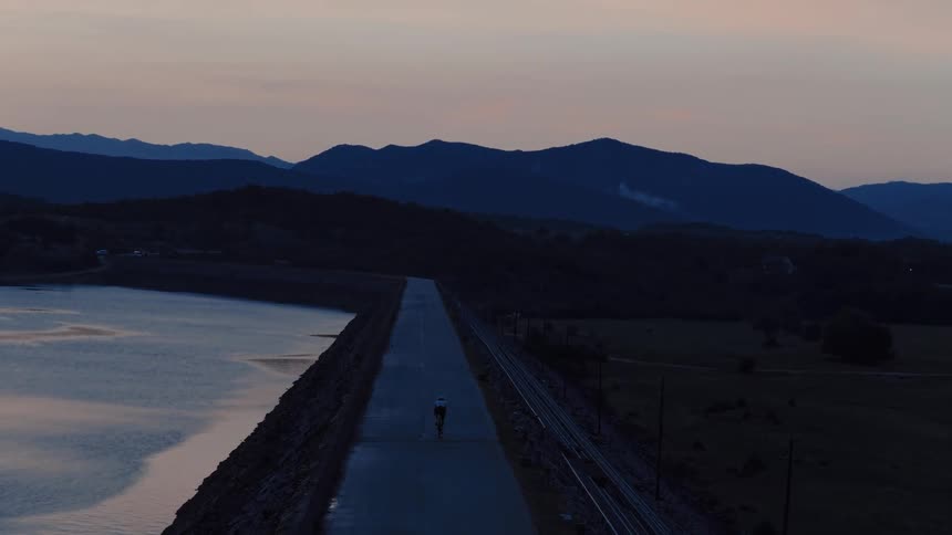 An aerial view of a solitary cyclist riding along a road on top of a dam next to a lake at dusk with mountains in the background