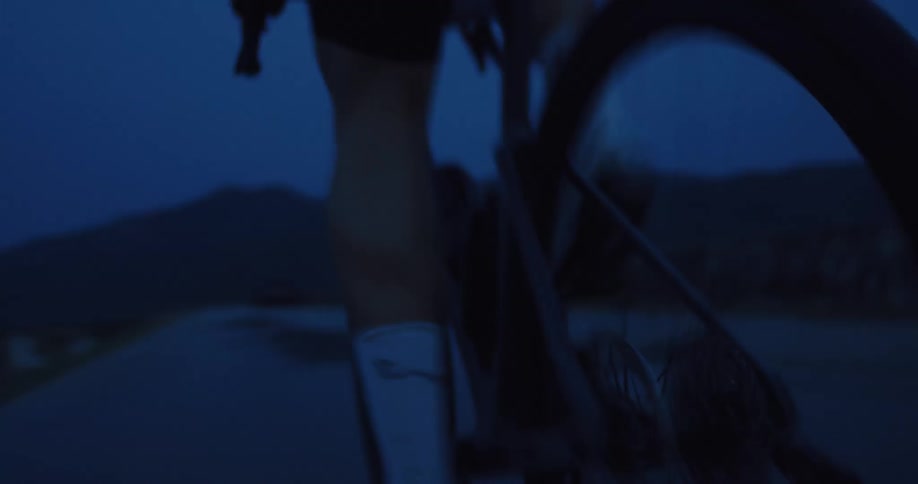 A low-angle close-up of a cyclist pedaling hard on a dark road at twilight, with the focus on the rear wheel and legs in motion