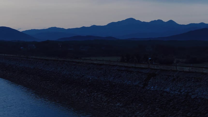 A lone cyclist rides across a dam road at dawn, with a tranquil reservoir on one side and a vast mountain range in the distance