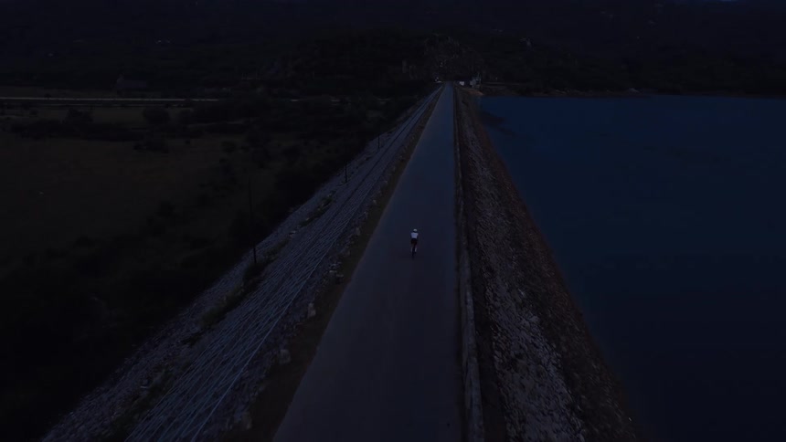 An aerial view shows a lone cyclist riding on a dam road beside a calm lake during the quiet blue hour
