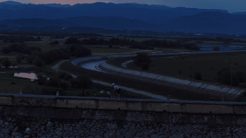 Aerial view of a lone cyclist riding along a dam road at dusk, with a winding river and mountains in the background