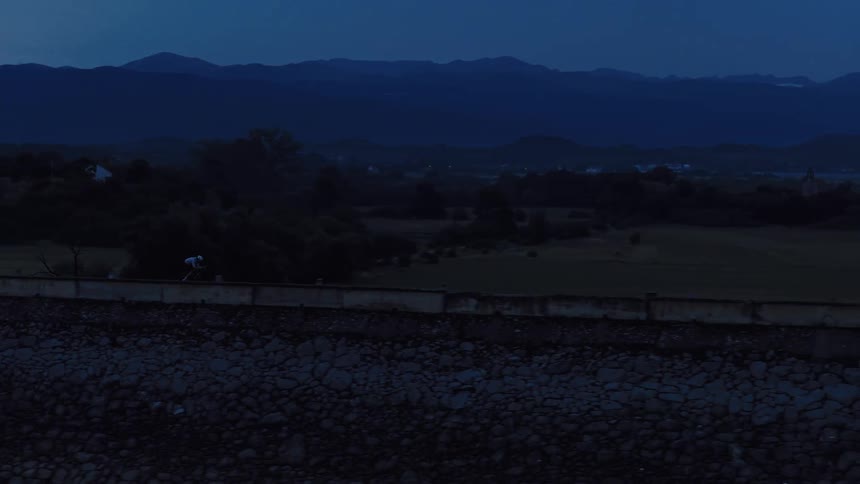 Aerial tracking shot of a cyclist riding across a dam during the blue hour with mountains in the background
