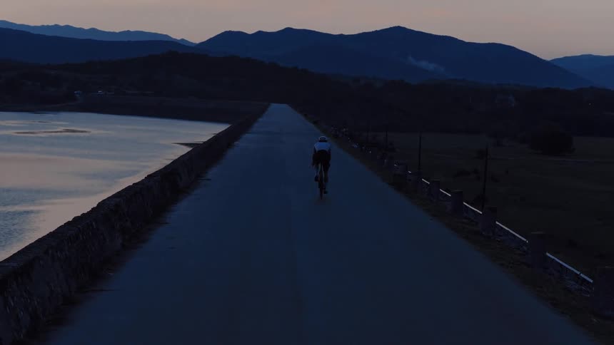 Aerial tracking shot follows a lone cyclist riding on a remote road across a dam at dusk with mountains on the horizon