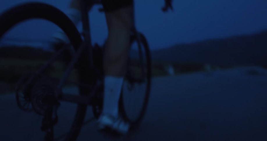 A close-up, low-angle shot captures the blur of a cyclist's legs as they pedal quickly along a road at dusk