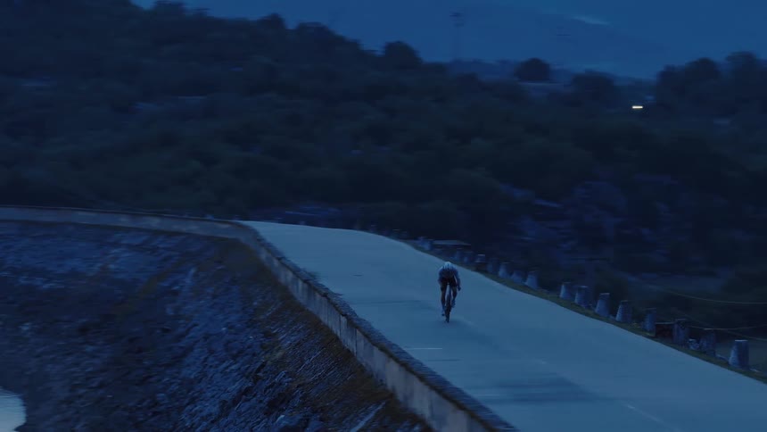 A lone cyclist rides along a curved road atop a dam in the dark, moody blue light of twilight