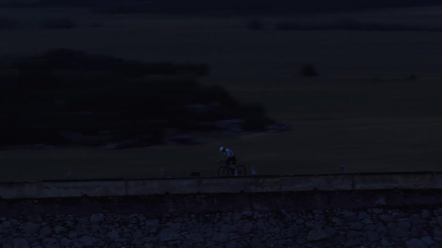 An aerial shot follows a lone cyclist riding along a quiet rural road on a dam at dusk
