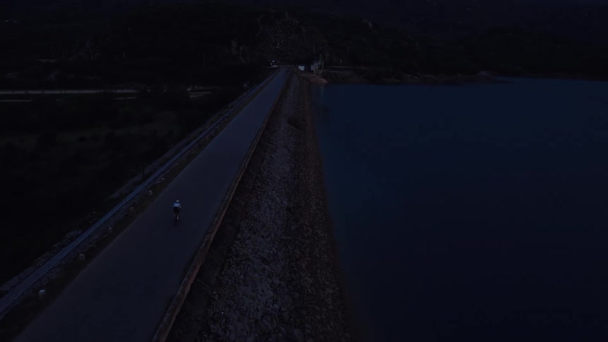 A lone cyclist rides across a remote dam road at twilight in this moody aerial shot
