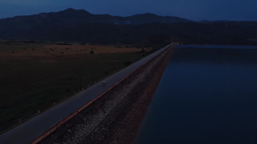 An aerial shot captures a lone cyclist riding down a long road on a dam next to a tranquil lake at dusk