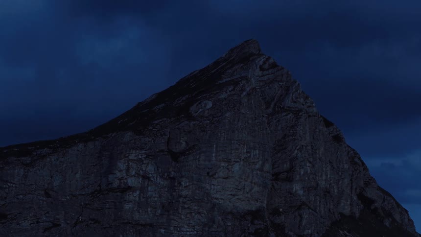 A rugged mountain peak stands silhouetted against a dark, stormy sky during the blue hour