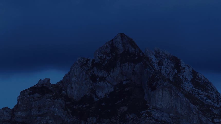 A slow zoom out reveals the jagged peaks of a mountain against a deep blue sky during the twilight hour