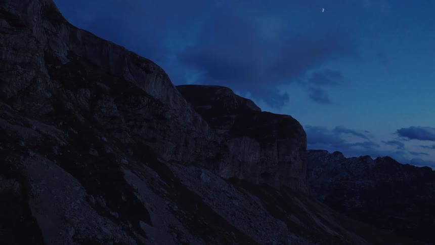 A crescent moon hangs in the deep blue sky over a dark, rocky mountain range at night